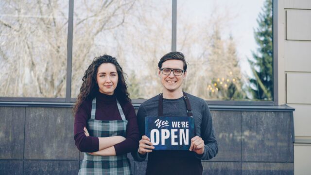 Dois donos de pequeno negocio segurando placa Yes We Are Open