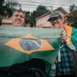 Dois homens sorridentes segurando a bandeira do Brasil ao ar livre em um bairro