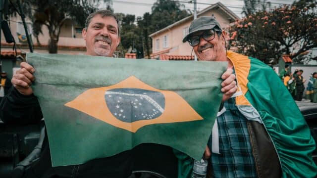 Dois homens sorridentes segurando a bandeira do Brasil ao ar livre em um bairro