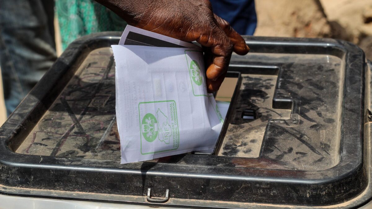 Mão colocando uma cédula de papel dentro de uma urna de votação transparente