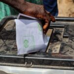 Mão colocando uma cédula de papel dentro de uma urna de votação transparente