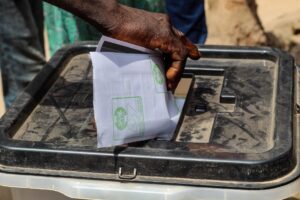 Mão colocando uma cédula de papel dentro de uma urna de votação transparente