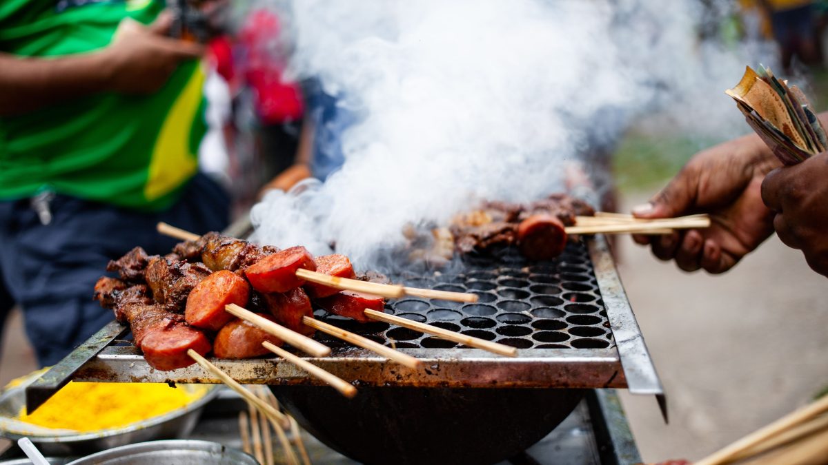 Espetos de carne grelhando sobre fogo, com fumaça subindo, em uma cena de churrasco de rua no Brasil