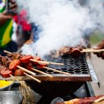 Espetos de carne grelhando sobre fogo, com fumaça subindo, em uma cena de churrasco de rua no Brasil