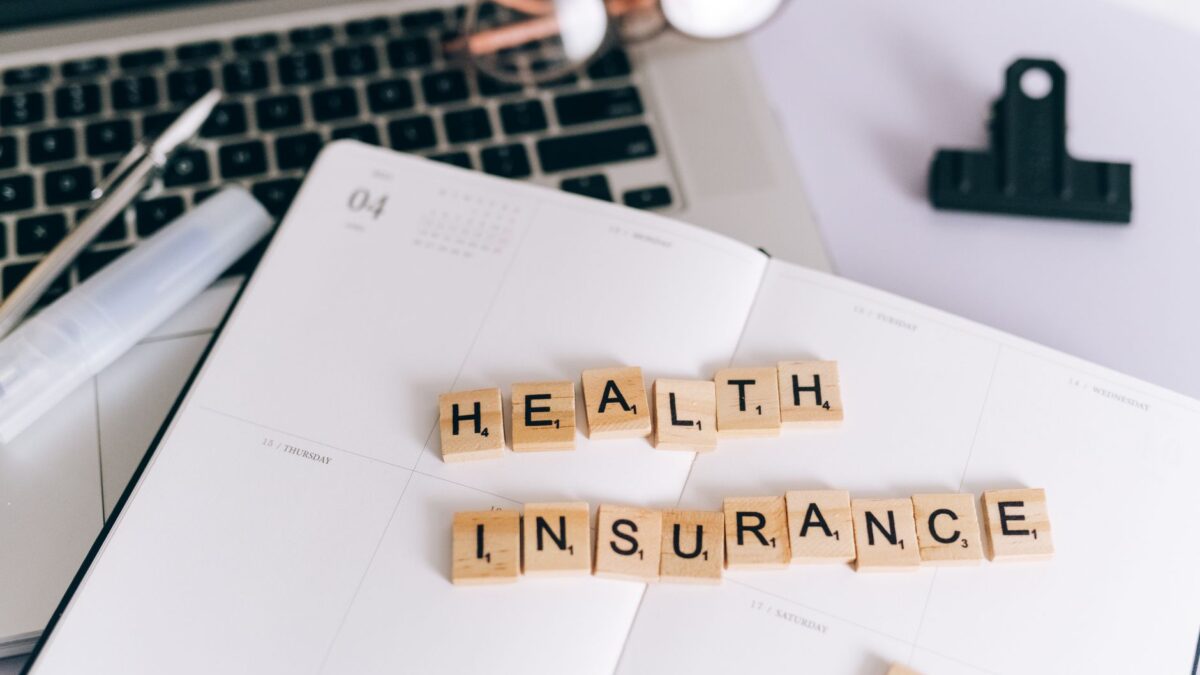 Peças de Scrabble formando as palavras "Health Insurance" sobre um planner, ao lado de um notebook